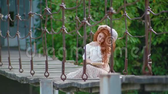 可爱的女孩浓密美丽的火红头发坐在河上的吊桥上看书视频的预览图