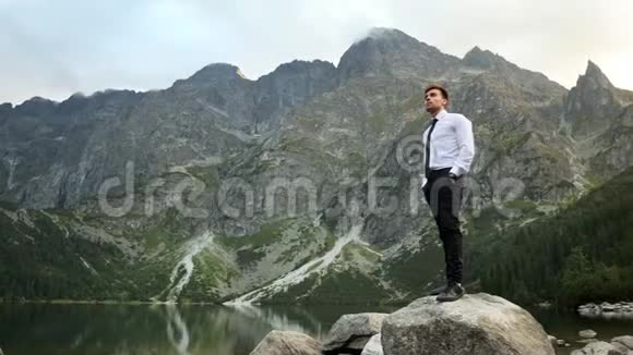 成功自信的商人在日落时分站在山上湖边的岩石上看着他的手表视频的预览图
