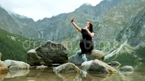 美丽的女游客坐在岩石湖附近的石头上用手机给朋友打视频电话视频的预览图