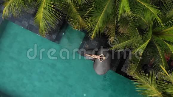 穿着米色比基尼和草帽的苗条年轻女子在豪华游泳池放松的空中无人机飞行景观假期视频的预览图