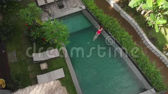 在热带花园别墅的游泳池里穿着红色比基尼的苗条年轻女子尽情放松享受日光浴视频的预览图