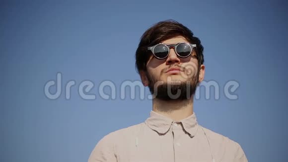 在蓝天背景上戴着圆太阳镜和衬衫的严肃胡须嬉皮士的肖像视频的预览图