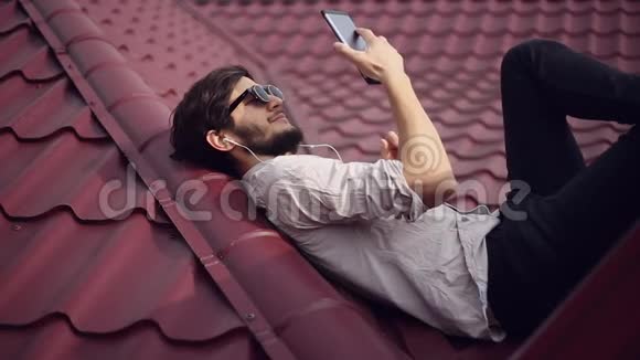 年轻快乐的家伙躺在红色的屋顶上用智能手机带耳机戴着圆形太阳镜和衬衫视频的预览图