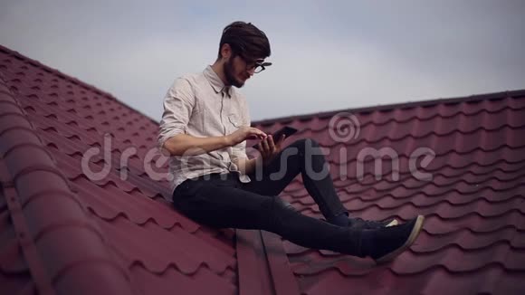 年轻开朗的嬉皮士坐在屋顶上手里拿着智能手机戴着圆圆的太阳镜和衬衫视频的预览图
