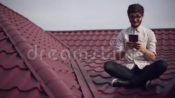 年轻开朗的嬉皮士坐在屋顶上手里拿着智能手机玩电子游戏戴着圆圆的太阳镜和衬衫视频的预览图