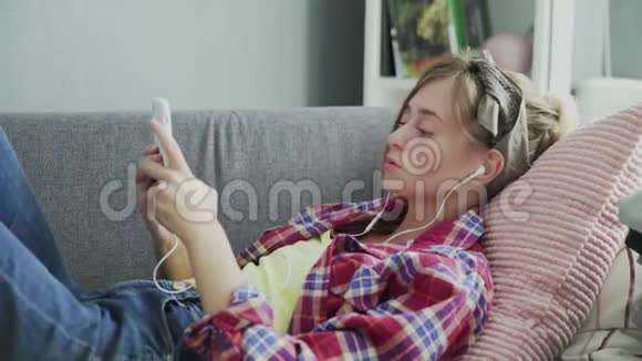 年轻的女人戴着耳机听着家里的音乐在沙发上视频的预览图