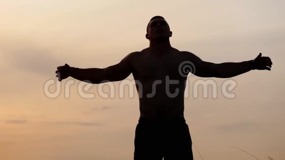 运动员的剪影裸露的躯干摆姿势显示他的肌肉和二头肌在日落时靠在天空体育保健视频的预览图