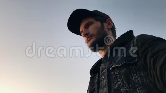 在天空背景上戴着黑色帽子和牛仔裤夹克的年轻留胡子的人的肖像视频的预览图