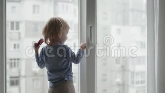 孩子在一扇开着的窗户附近有危险小男孩站在窗台上手里拿着玩具车向外看视频的预览图