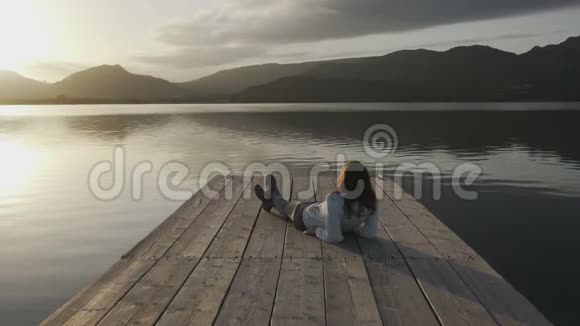 一位从背后看到长发的年轻女子在夕阳下看湖她躺在一个古老的木墩上视频的预览图