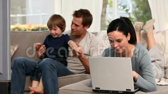 女人用笔记本电脑而她的男孩和丈夫看电视视频的预览图