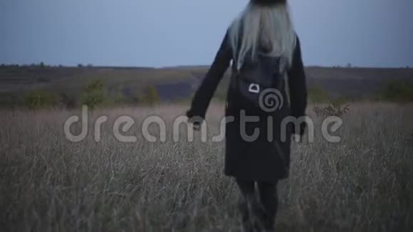 背面的年轻金发女孩穿着黑色戴着帽子外套和背包在干燥的田野里行走视频的预览图