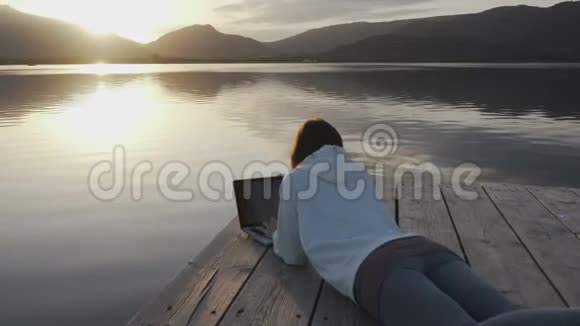 网络自由职业选择概念一位年轻女性在日落时分躺在湖边的码头上的笔记本电脑上工作视频的预览图