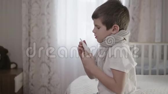 生病的孩子戴着温暖的围巾鼻塞在流感期间用滴药私下坐在床上视频的预览图