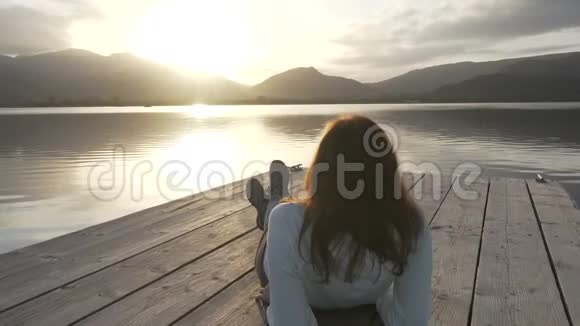 一位从背后看到长发的年轻女子在夕阳下看湖她躺在一个古老的木墩上视频的预览图
