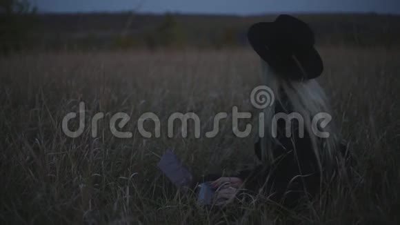 金发女孩的肖像戴着黑色帽子和外套坐在田野里的干草上腿上有笔记本电脑视频的预览图