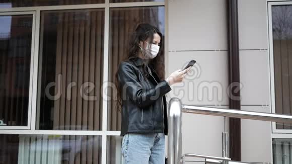 冠状病毒保护一位戴着医用防护面罩的年轻女子站在一条城市街道上手里拿着智能手机视频的预览图