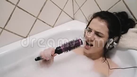 年轻女孩躺在泡沫浴缸里听着耳机里的音乐用梳子唱歌视频的预览图