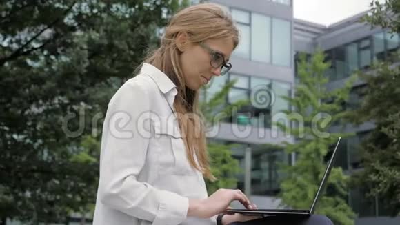 年轻的商务女性在绿色公园的办公楼旁使用便携式电脑视频的预览图
