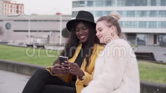 一个金发的北欧女孩和一个黑人非裔美国女孩坐在城里的长凳上用手机视频的预览图