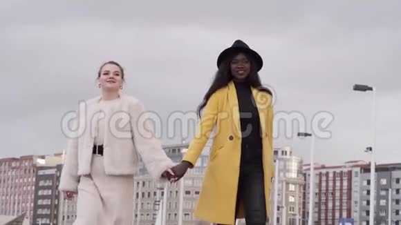 两个女孩金发女孩和一个黑人非裔美国女孩携手穿过城市视频的预览图