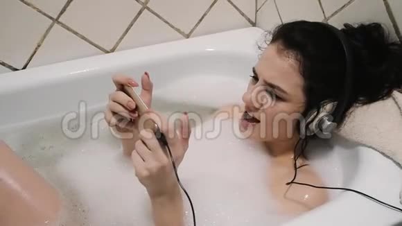 年轻女孩用防水智能手机耳机听音乐在泡泡浴中唱歌视频的预览图