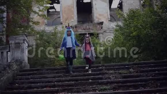 两个兴高采烈的小女孩穿着五颜六色的独角兽服装蓝色的头发和绿色的万圣节桶走在废弃的楼梯上视频的预览图