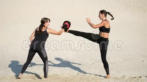 两个穿着黑色健身服的年轻女子在一个荒无人烟的海滩上一对一对地练习踢腿视频的预览图