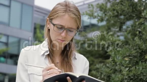 年轻的学生女孩戴着眼镜坐在户外长凳上写笔记视频的预览图