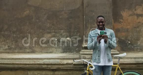 兴奋的美国黑人年轻人用他的智能手机玩在线游戏同时站在前面的老建筑墙附近视频的预览图