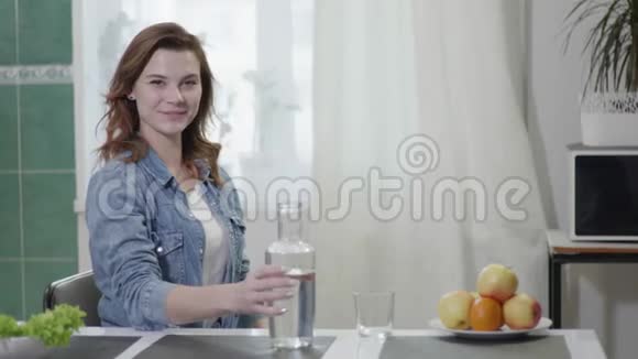 迷人的微笑女孩把干净的水倒进杯子里一边喝饮料一边看着相机一边坐在厨房的桌子上视频的预览图