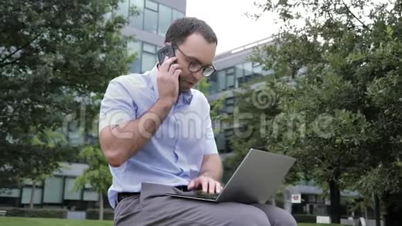 在公园户外工作的商人使用笔记本电脑和移动电话视频的预览图