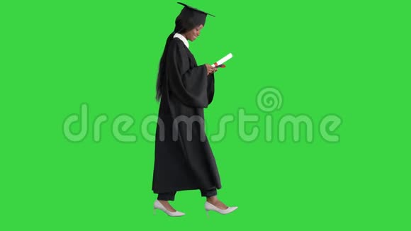 年轻的美国黑人女学生身穿黑色毕业礼服持有文凭在绿色上使用手机视频的预览图