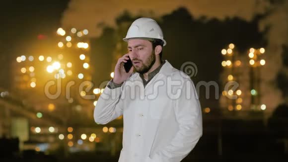 年轻愤怒的工程师戴着头盔穿着白色长袍站在炼油厂的电话里大喊大叫视频的预览图
