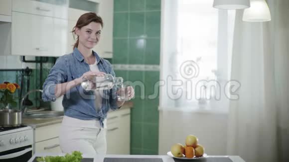 年轻可爱的女人把水倒进玻璃里然后喝着它看着相机微笑着站在厨房的桌子上视频的预览图
