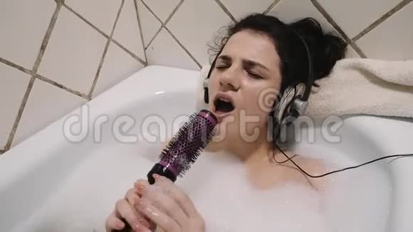 年轻女孩躺在泡沫浴缸里听着耳机里的音乐用梳子唱歌视频的预览图