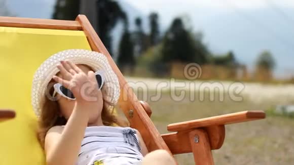旅游女婴坐在甲板椅上挥舞着手戴着太阳镜和帽子的小女孩吃糖果和视频的预览图