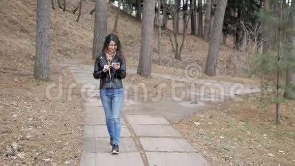 美丽的年轻开朗的女人穿着皮夹克走在秋天公园用手机说话视频的预览图