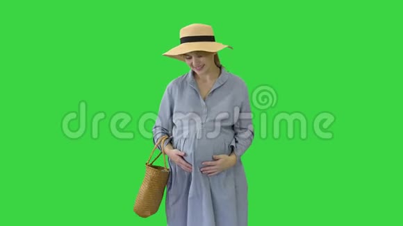 美丽的孕妇穿着夏天的衣服戴着帽子摸着她的腹部在绿色屏幕上Chroma键视频的预览图