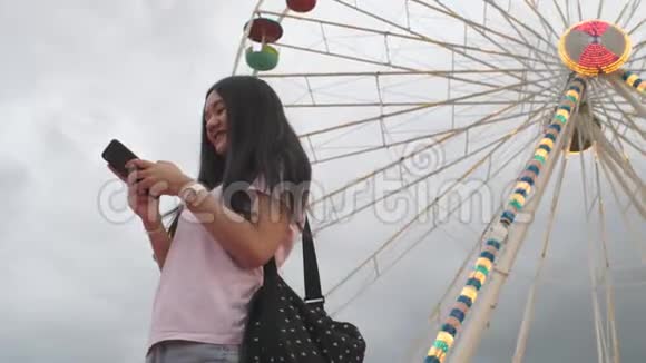 快乐美女亚洲女人看智能手机拍自拍照片给朋友游乐园用摩天轮骑视频的预览图