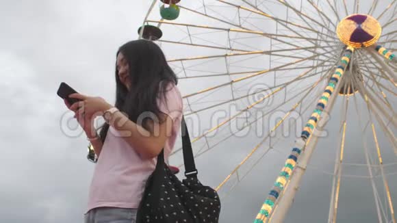 快乐美女亚洲女人看智能手机拍自拍照片给朋友游乐园用摩天轮骑视频的预览图