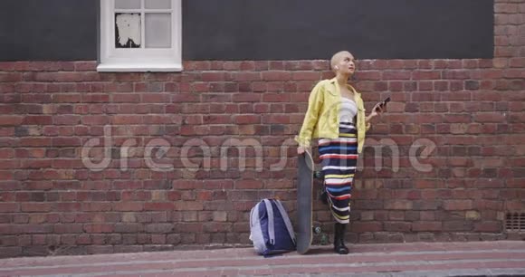 混合种族的女人靠在砖墙上她的滑板和她的手机视频的预览图