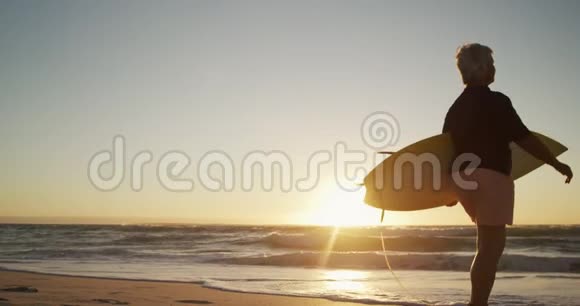 从侧面看老女人在海滩上拿着冲浪板走路视频的预览图
