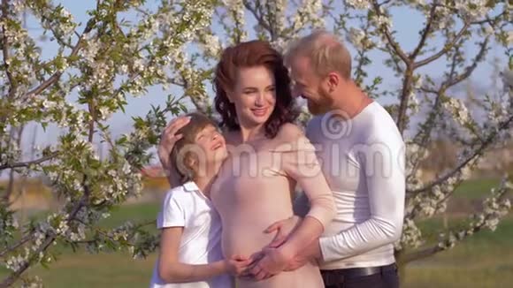 快乐的德国家庭微笑的男人与孕妇和十几岁的儿子拥抱在开花的春天花园靠近果树视频的预览图