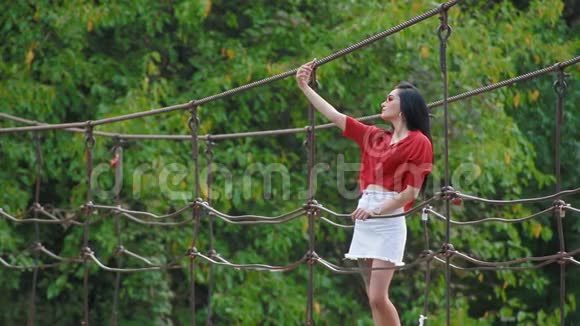 穿着鲜红衬衫的女人站在河上的吊桥上在手机上自拍视频的预览图