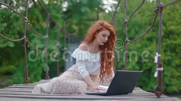 年轻的商务女性浓密美丽的火红头发坐在河上的吊桥上手提电脑工作视频的预览图