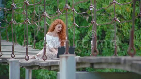 年轻的商务女性浓密美丽的火红头发坐在河上的吊桥上手提电脑工作视频的预览图