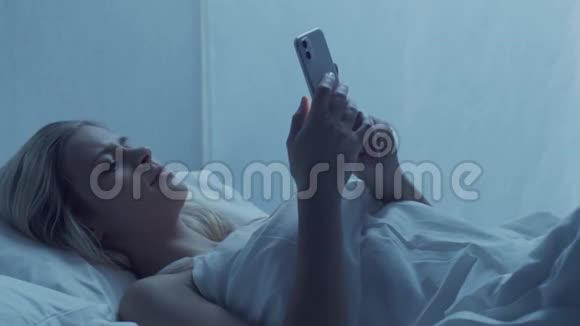 年轻的女人晚上躺在床上拿着智能手机美丽的金发女孩休息卧室里的暮色视频的预览图