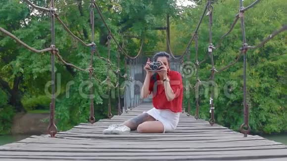 年轻的黑发女子带着旧相机坐在河上的吊桥上直接对着镜头拍照视频的预览图
