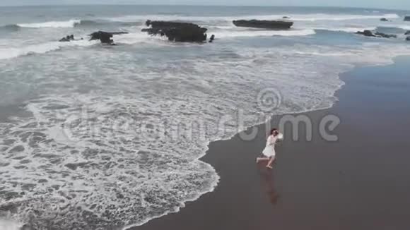鸟瞰美丽的女人穿着白色连衣裙在海岸线上奔跑在沙滩上享受夏日的黑沙假期视频的预览图